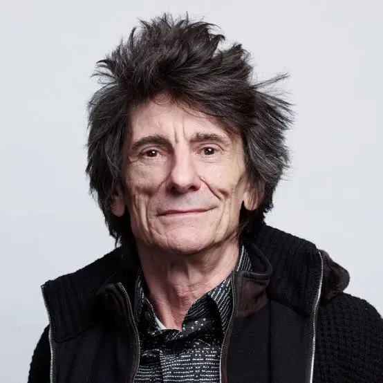 Retrato de primer plano del guitarrista Ronnie Wood de The Rolling Stones sonriendo sobre fondo gris.