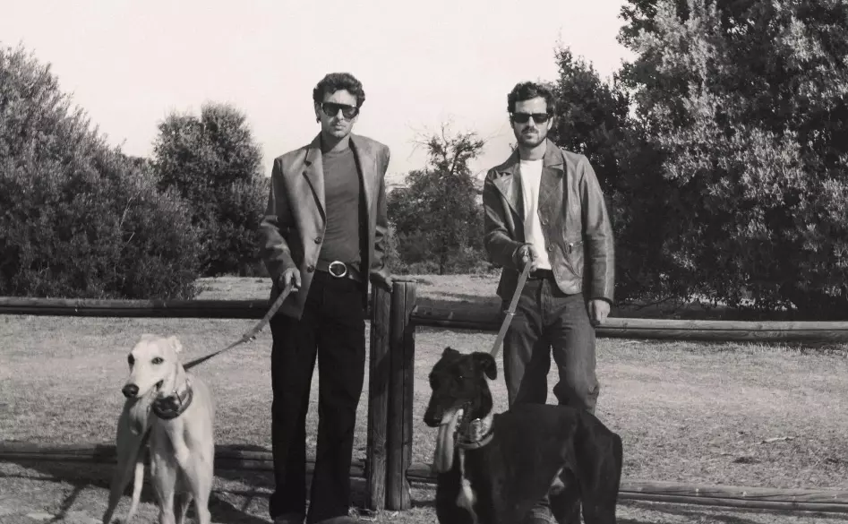 Willy Bárcenas y Antón Carreño de Taburete posando en el campo con gafas de sol y chaquetas de cuero junto a dos perros galgos para la promoción de su gira 2026.